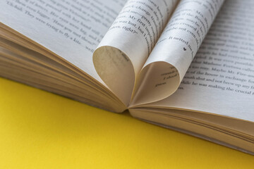 Love of books, reading. An open book with curled pages of books in the shape of a heart on a yellow background. Place to copy space.