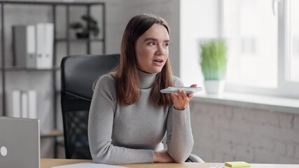 Smiling young woman recording voice message on smartphone. Beautiful girl using virtual assistant app sitting at home office. Voice recognition, audio messages, using translator on mobile phone