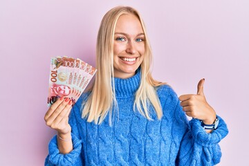 Young blonde girl holding 100 new zealand dollars banknote smiling happy and positive, thumb up doing excellent and approval sign