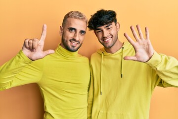 Homosexual gay couple standing together wearing yellow clothes showing and pointing up with fingers number seven while smiling confident and happy.