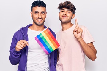 Young gay couple holding rainbow lgbtq flag smiling with an idea or question pointing finger with happy face, number one