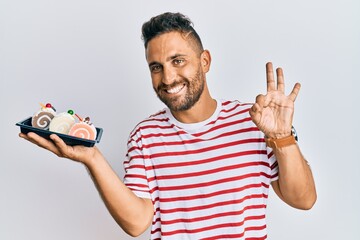 Handsome man with beard holding cake sweets doing ok sign with fingers, smiling friendly gesturing excellent symbol