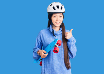 Young beautiful chinese woman wearing security helmet holding skate smiling happy and positive, thumb up doing excellent and approval sign