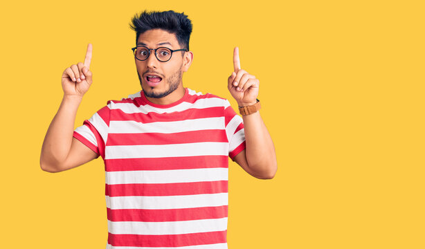 Handsome latin american young man wearing casual clothes and glasses smiling amazed and surprised and pointing up with fingers and raised arms.