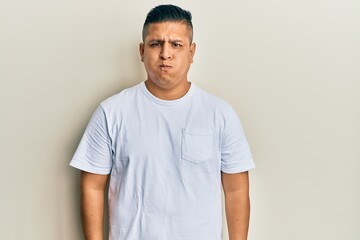 Young latin man wearing casual white t shirt puffing cheeks with funny face. mouth inflated with air, crazy expression.