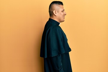 Young latin priest man standing over yellow background looking to side, relax profile pose with...