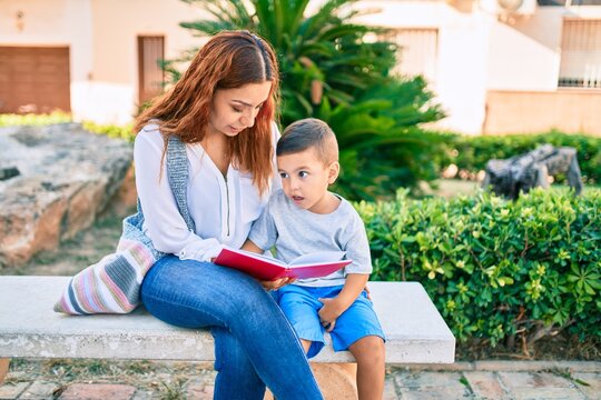 Adorable latin mother and son sitting on the bench and reading book at the park.