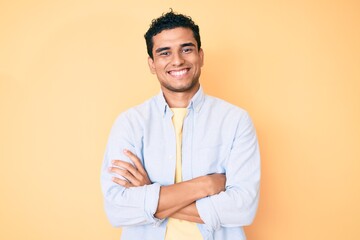 Young handsome hispanic man standing over yellow background happy face smiling with crossed arms...