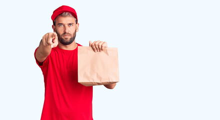 Naklejka premium Young handsome blond man holding delivery paper bag pointing with finger to the camera and to you, confident gesture looking serious