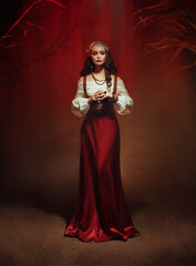 Young beautiful gypsy woman stands in a dark room. Long black flowing hair, rose hairpin. Red...