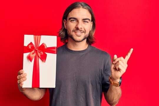 Young handsome man holding gift smiling happy pointing with hand and finger to the side