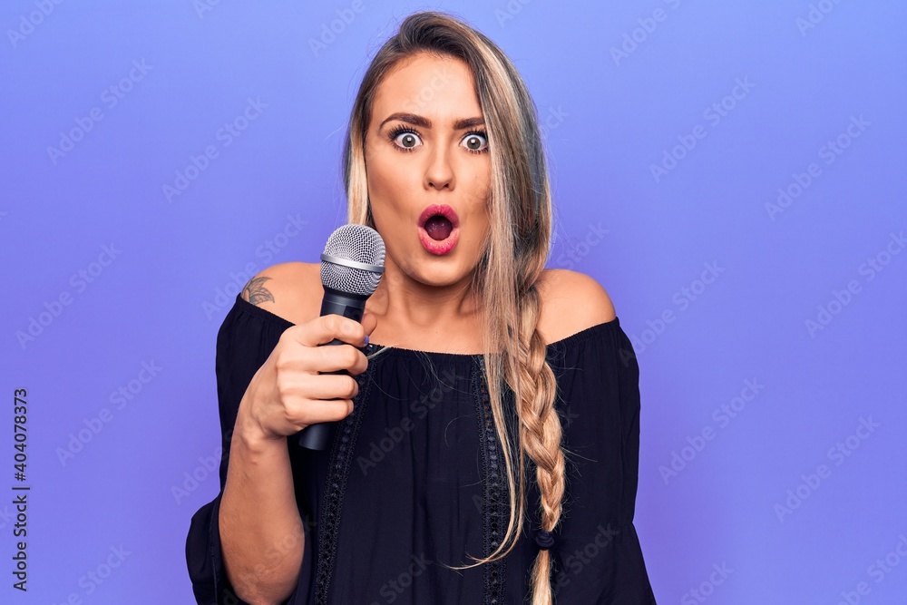 Poster young beautiful blonde singer woman singing using microphone over purple background scared and amaze