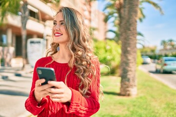 Young blonde girl smiling happy using smartphone standing at the park