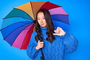 Young beautiful chinese girl holding colorful umbrella with angry face, negative sign showing dislike with thumbs down, rejection concept