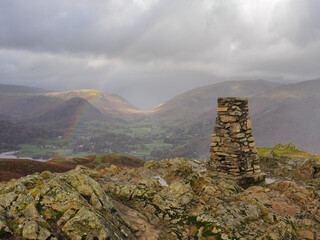 Triangulation point at the top of Loughrigg Fell overlooking Grasmere with a rainbow arcing across...