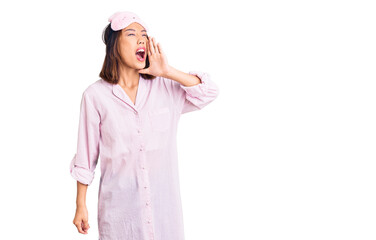 Fototapeta na wymiar Young beautiful chinese girl wearing sleep mask and pajama shouting and screaming loud to side with hand on mouth. communication concept.