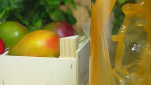 Close-up of mango juice poured in a jug next to the box full of mango on the background of greenery in slow motion