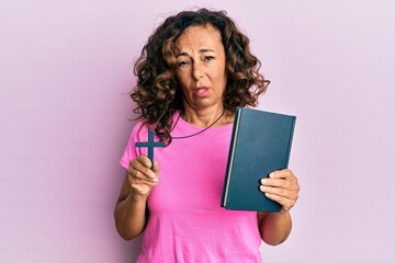 Middle age hispanic woman holding bible and christian cross clueless and confused expression. doubt...