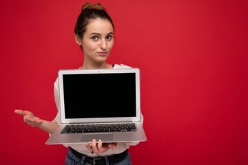 Close-up portrait of Beautiful smiling happy young woman holding computer laptop looking at camera having questions wearing casual smart clothes isolated over red wall background