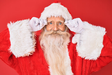Old senior man with grey hair and long beard wearing santa claus costume holding glasses smiling with a happy and cool smile on face. showing teeth.