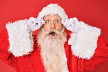 Old senior man with grey hair and long beard wearing santa claus costume holding glasses afraid and...