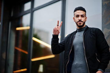 Fototapeta premium Portrait of stylish arab beard man wear grey turtleneck and black jaket. Arabian model guy show two fingers.