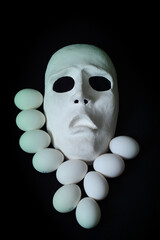 A white scary theater mask lies next to the chicken eggs