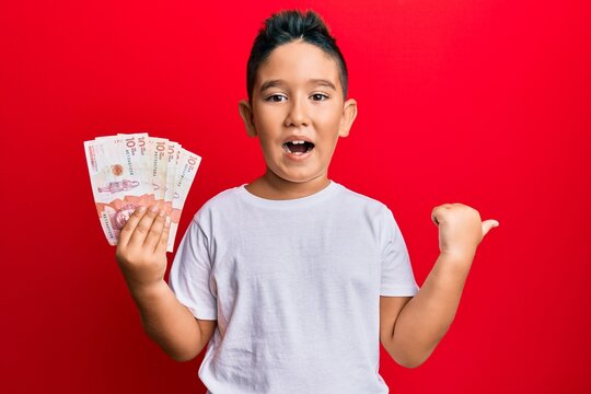 Little Boy Hispanic Kid Holding 10 Colombian Pesos Banknotes Pointing Thumb Up To The Side Smiling Happy With Open Mouth