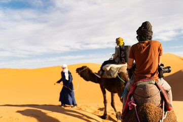 (Selective focus) Stunning view of two people riding camels on the sand dunes in Merzouga, Morocco. Merzouga is a small village in southeastern Morocco.