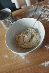 White bowl with dough and spoon in it, on the wooden table in the kitchen. The process of mixing ingredients for pastry. Closeup, selective focus.