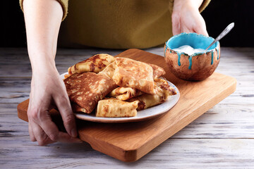 Homemade crepes, tasty thin pancakes on plate with a bowl of sour cream on old wooden plank in female hands