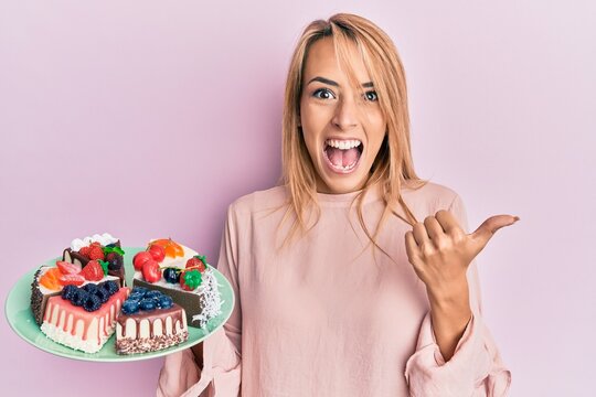 Beautiful blonde woman holding cake slices pointing thumb up to the side smiling happy with open mouth