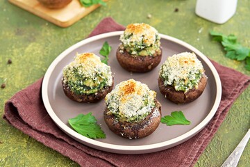 Baked brown mushrooms stuffed with spinach and cheese, with breadcrumbs and cheese crust on a brown plate on a green concrete background. Champignon recipes.