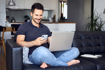 Handsome man sitting with laptop at home using mobile phone