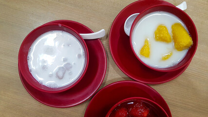 Top view of Thai dessert with strawberry in syrup, banana in coconut milk and taro Dumplings in coconut cream in red bowl on wooden table or background. Sweet Asian food.