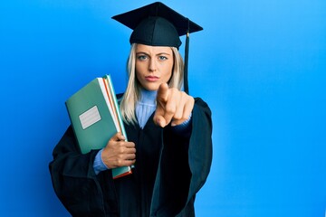 Beautiful blonde woman wearing graduation cap and ceremony robe holding books pointing with finger to the camera and to you, confident gesture looking serious