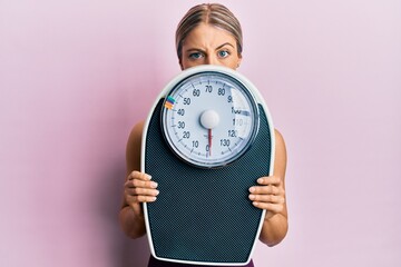 Beautiful blonde woman holding weight machine to balance weight loss skeptic and nervous, frowning...