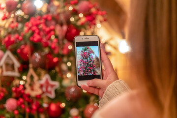 Close-up view of woman in white sweater photographing a Christmas tree using her smart phone. Blurred Christmas tree with garland lights in the background. - Powered by Adobe