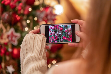 Close-up view of woman in white sweater photographing a Christmas tree using her smart phone. Blurred Christmas tree with garland lights in the background.