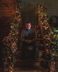 Obraz na płótnie Canvas Portrait of caucasian redhead man sitting on wooden staircase decorated for Christmas and New Year. Smiling man wears casual clothes: jeans and a gray sweater. Christmas home celebration theme.