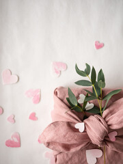 Gift wrapped in pink linen decorated with eucalyptus on a background of paper pink hearts on a white background in the morning light