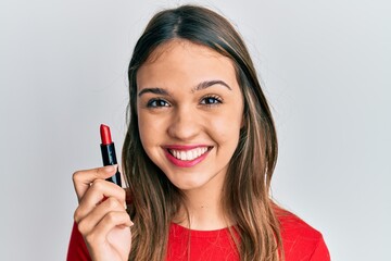 Young brunette woman holding red lipstick looking positive and happy standing and smiling with a confident smile showing teeth