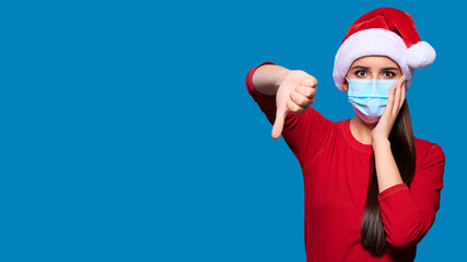 Excited and shocked woman wearing Santa hat and face protective medical mask showing thumb down gesture, blue isolated background.