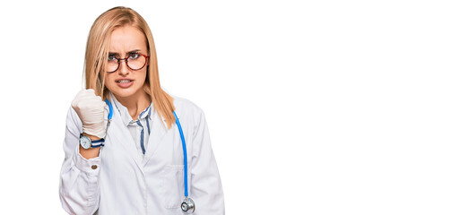 Beautiful caucasian woman wearing doctor uniform and stethoscope angry and mad raising fist frustrated and furious while shouting with anger. rage and aggressive concept.