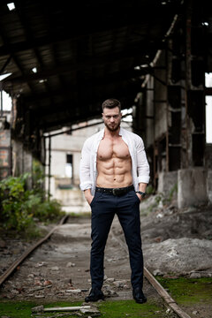 Very muscular man with unbuttoned white shirt and naked torso poses on abandoned storehouse background.