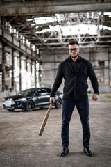Full lenght portrait of a cool brutal guy with baseball bat in hands and modern car on the background. Total black outfit with sunglasses.