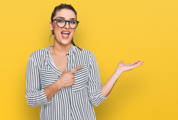 Young caucasian woman wearing business shirt and glasses amazed and smiling to the camera while presenting with hand and pointing with finger.