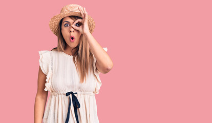 Young beautiful woman wearing summer hat and t-shirt doing ok gesture shocked with surprised face, eye looking through fingers. unbelieving expression.