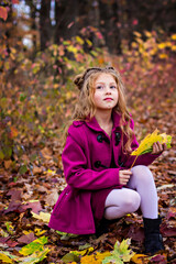 Beautiful little cute blonde girl in a pink coat stands near the wall with autumn red leaves.