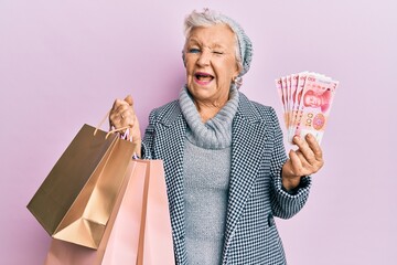 Senior grey-haired woman holding shopping bags and israel shekels banknotes winking looking at the...
